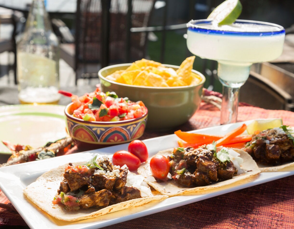 A close-up image of a hand squeezing lime onto a taco filled with seasoned shredded pork, showcasing traditional Mexican street food served on a wooden plate and margarita